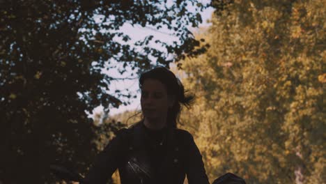 Pretty-smiling-European-young-woman-driving-a-motorbike-wearing-leather-jacket-in-forest-with-vibrant,-colorful-golden-autumn-leaves-on-sunny-day-2