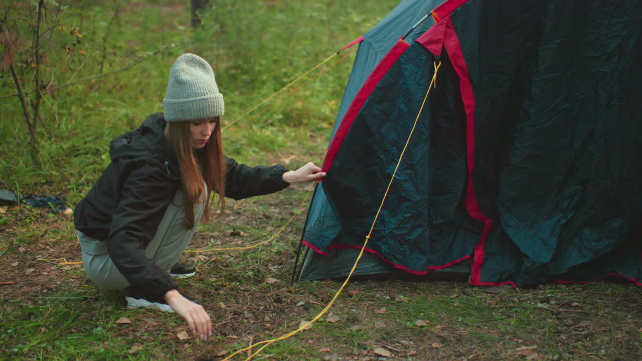 Lady squats beside forest tent carefully removing rope from tent pole, dressed in beanie and casual wear, surrounded by green grass and woodland environment, focused on camping task