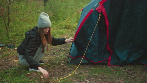 lady squats beside forest tent carefully removing rope from tent pole, dressed in beanie and casual wear, surrounded by green grass and woodland environment, focused on camping task