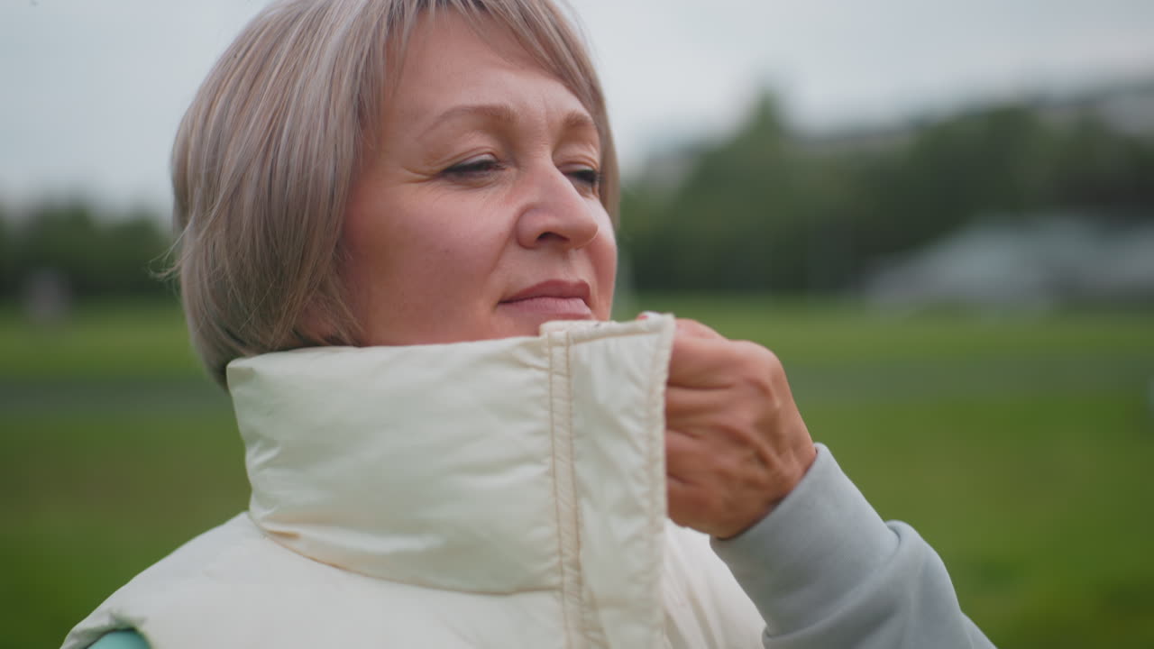 Close up of trainer wearing mint green hoodie and gray pants carefully zipping puffy sleeveless coat from bottom to top outdoors on grassy field, preparing for outdoor activity on cool cloudy day