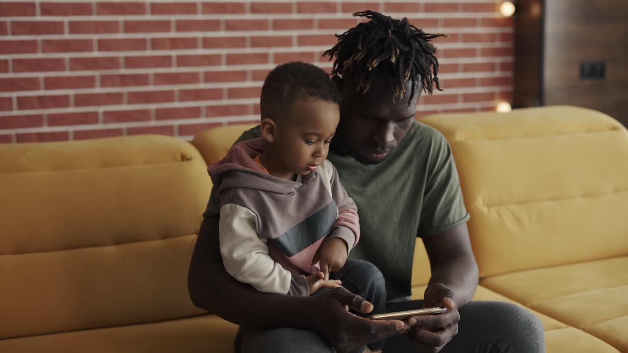Portrait of african american father sitting with son on sofa using smartphone