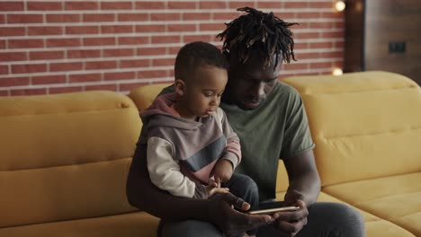 portrait of african american father sitting with son on sofa using smartphone