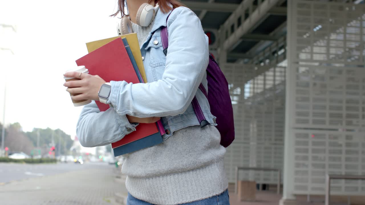 Úvodní záběr studentky objímající sešity a hrnek kávy na autobusové zastávce, upíjející a čekající na příjezd autobusu.