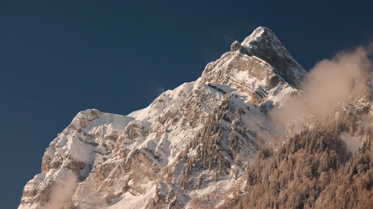 A telephoto shot of a snow-covered alpine mountain peak under a clear blue sky. The static tripod footage captures calm winter scenery and natural mountain textures in daylight
