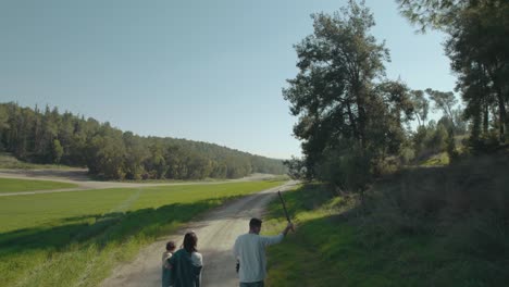 Mother,-father,-little-girl-and-vizsla-dog-walk-in-a-park-near-an-open-field,-the-mother-carries-the-girl-and-the-father-throws-a-stick-to-the-dog---push-in-shot-from-the-back