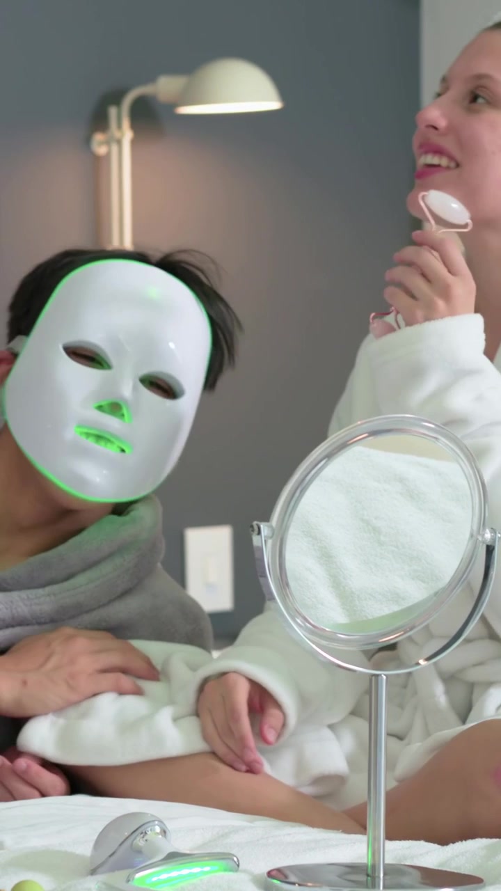 Video verticale: Donna in accappatoio che entra e rimuove una maschera LED da un uomo a letto, usando un rullo per la cura personale.