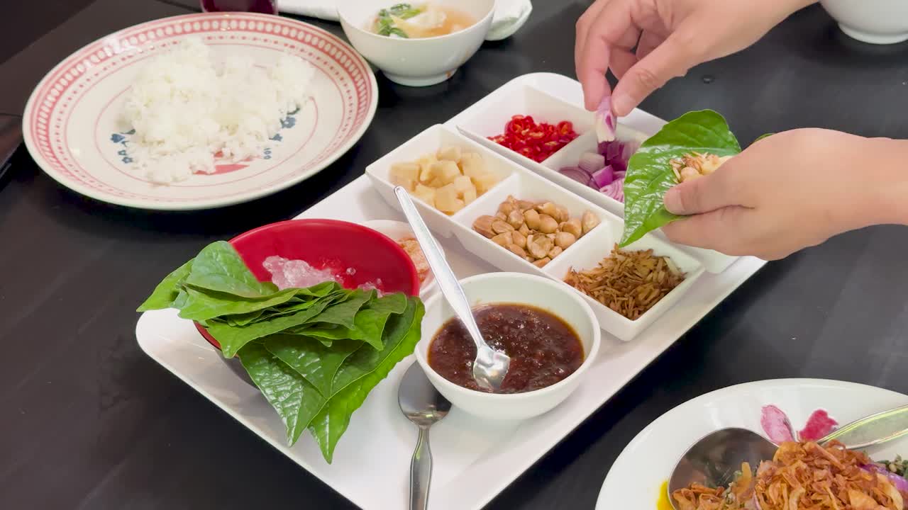 Hands assemble Miang Kham savory leaf wraps with assorted fillings and sauce on a white tray, under bright indoor lighting, viewed from above