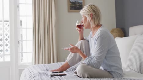 happy senior caucasian woman sitting on bed in bedroom, using smartphone and drinking coffee