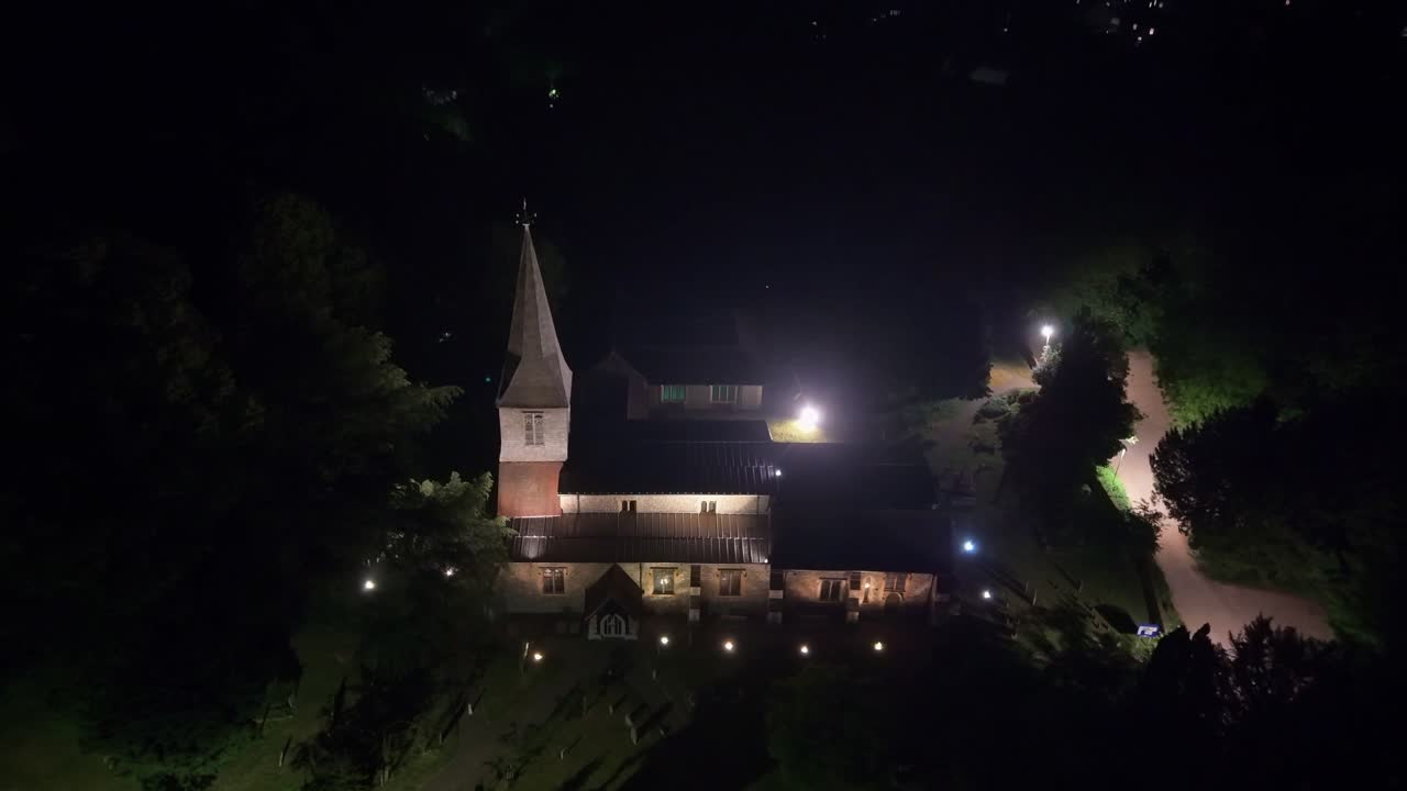 Aerial drone pull back reveals a glowing illuminated St Stephen's Church from above, with the twinkling city lights of St Albans in the background during the peaceful evening hou
