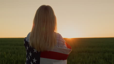 a woman with an american flag on her shoulders watches the sun go down over a field of wheat.