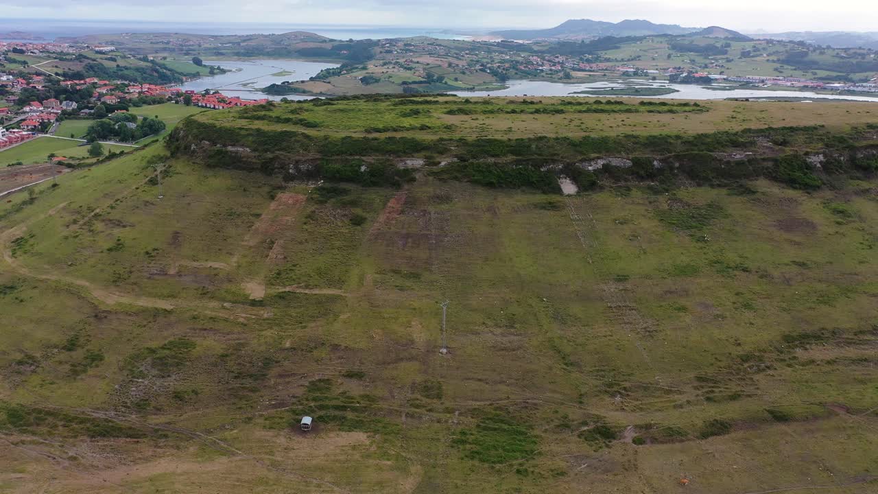 vuelo lateral que muestra los restos de un asentamiento paleolítico en una zona verde y rodeado de una zona urbana y montañas con la desembocadura del río al mar en cantabria, españa
