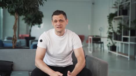 portrait-of-a-young-man-in-a-white-T-shirt-against-the-backdrop-of-a-business-center,-sitting-on-a-sofa,-folding,-talking,-looking-into-the-camera,-and-gesticulating