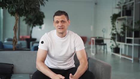 portrait of a young man in a white t-shirt against the backdrop of a business center, sitting on a sofa, folding, talking, looking into the camera, and gesticulating
