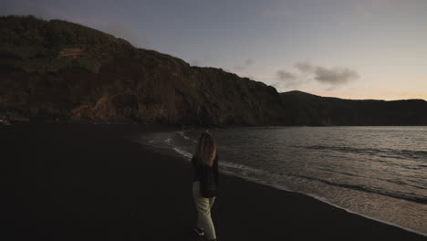 Woman-walks-on-beach-by-cliffs-at-the-Azores-at-dusk,-wide-handheld