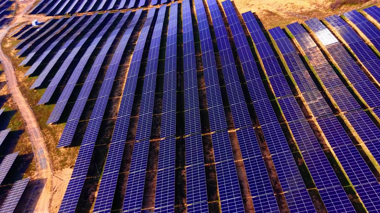 File diagonali di pannelli solari con riflessi illuminati dal sole, Bulgaria. Vista aerea obliqua di moduli fotovoltaici blu che luccicano sotto la luce del giorno con piste di accesso sterrate che attraversano l'inquadratura