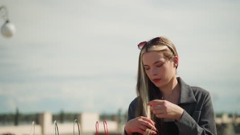 mujer con ropa gris ajusta su cabello mientras posa al aire libre, la luz del sol se refleja en ella, bolsas de compras de colores están a su lado mientras toma una postura relajada, en un día soleado y brillante