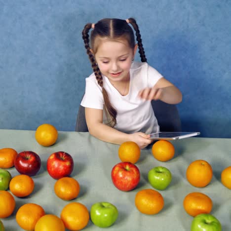 happy child with many organic fruit in the kitchen. little girl playing on digital tablet and counts fresh fruits on the table.