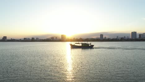 Un-Barco-De-Pesca-Al-Atardecer-Con-El-Horizonte-De-La-Ciudad-De-Vietnam-En-Segundo-Plano