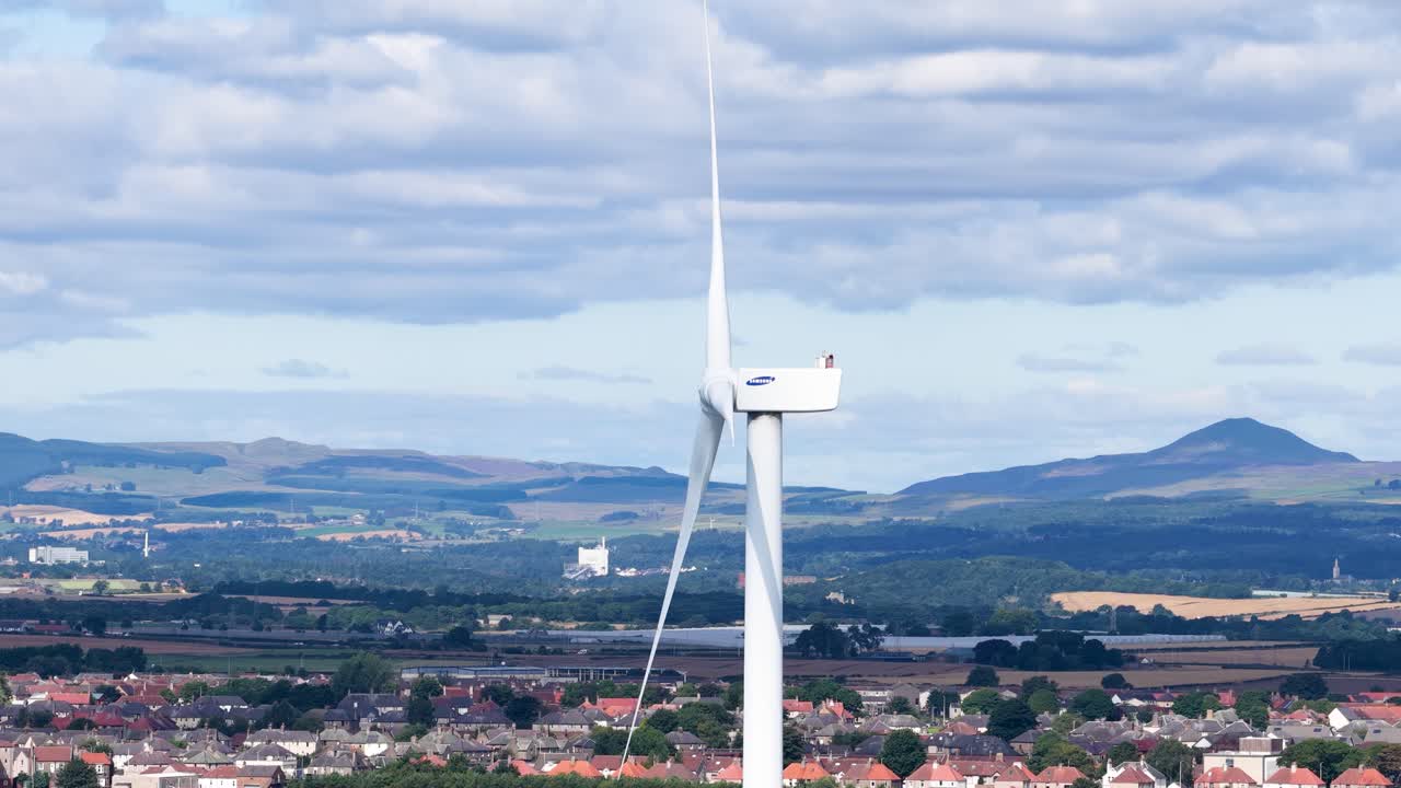 Large wind turbine spins steadily above Dundee rooftops, under bright daylight and partly cloudy skies