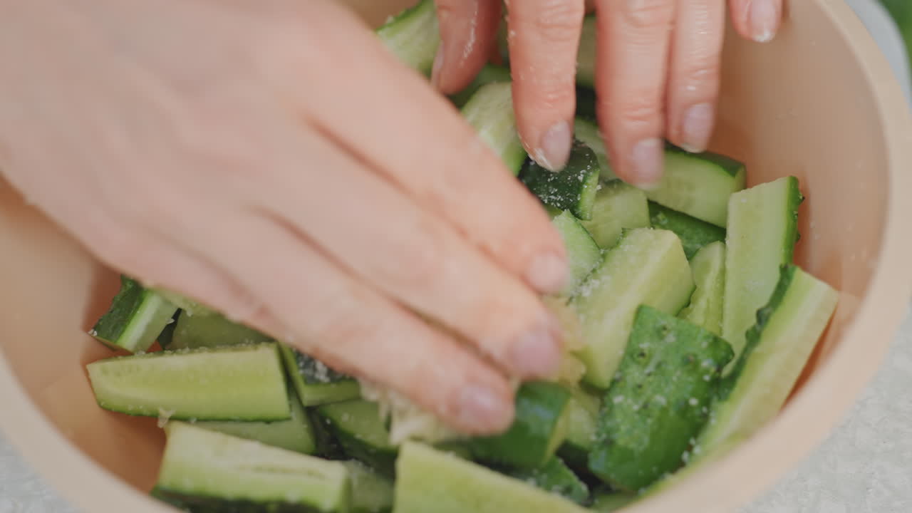 Primo piano di mani che mescolano pezzi di cetriolo in una ciotola, salando e rigirando per uno spuntino croccante da campeggio, preparazione tattile intima su un tavolo portatile con fette verdi fresche che brillano di condimento.