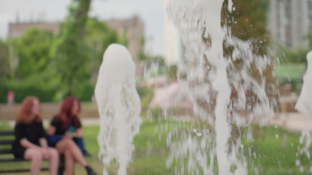 Water fountain jet in focus with blurred parents and kids in park background, summer greenery framing casual family outing, dynamic droplets catching light, quiet observation of spray