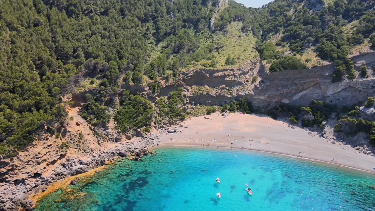 Experience the stunning landscape at Coll Baix beach in Puerto de Alcudia, Mallorca. Relax on the sandy shore, enjoy the clear blue waters, and soak in the sun amidst lush hills.