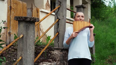 A-young-woman-standing-near-an-old-house-and-playing-the-pan-flute