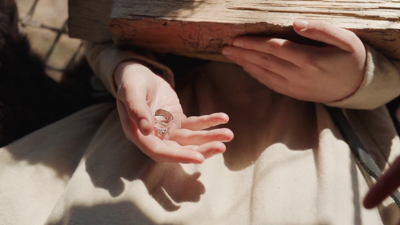 Un primo piano mostra mani che offrono e trasferiscono una minuscola conchiglia o un seme tra i palmi accanto a un libro aperto, con luce solare calda, ombre morbide, un tocco delicato e un'atmosfera di lettura tranquilla e concentrata.