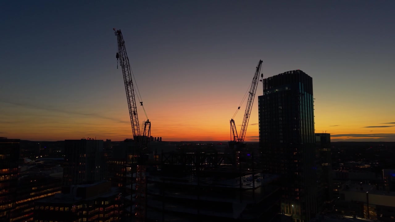 Aerial view of construction cranes and a skyscraper silhouetted against a vibrant sunset sky in Kendall Square, Cambridge, Massachusetts, United States.