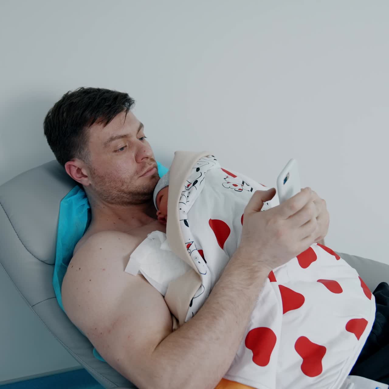 Caucasian man holds his newborn child on the chest. Father is looking at phone while baby is resting