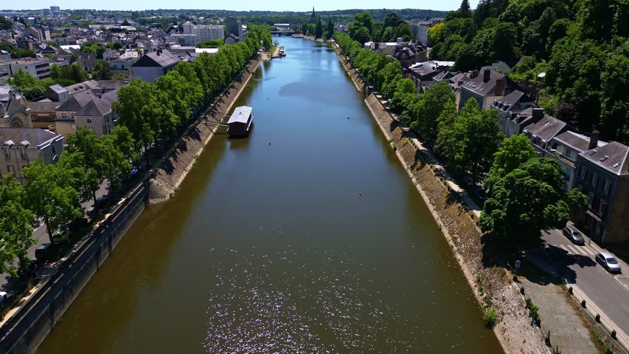Drone flight over Laval reveals the Mayenne River, riverside trees, city center, and Saint-Julien Chapel in sunlight. France