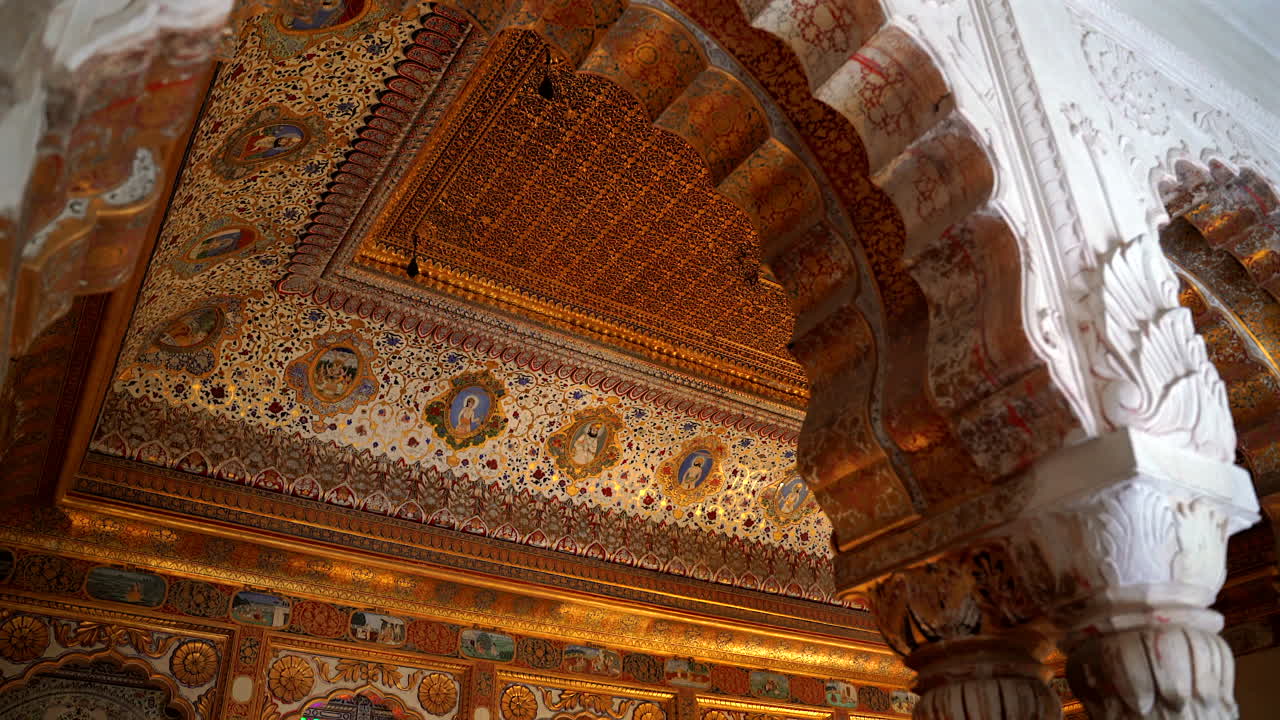 Un'inquadratura statica dal basso dell'opulento interno del Phool Mahal, che mostra il soffitto in foglia d'oro finemente dipinto, gli archi decorativi e i ritratti reali nel Forte di Mehrangarh, Jodhpur, India