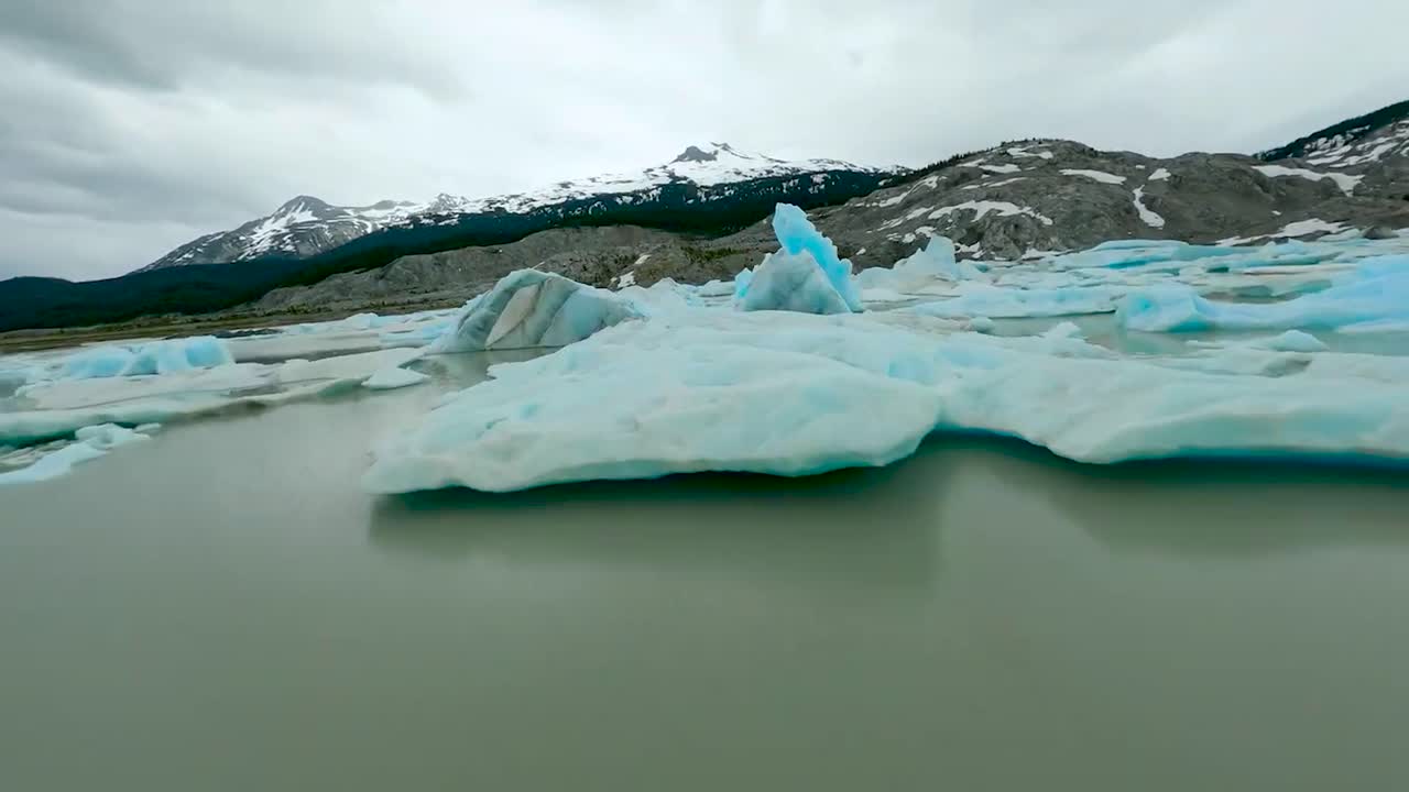FPV aerial drone flying over between and through icy blue and white glaciers in Iceland or Greenland arctic water during cloudy weather while mountains and rocky terrain is visible in the background.