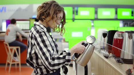 side view of a young cheerful positive attractive woman in plaid shirt choosing electric kettle in household appliances store