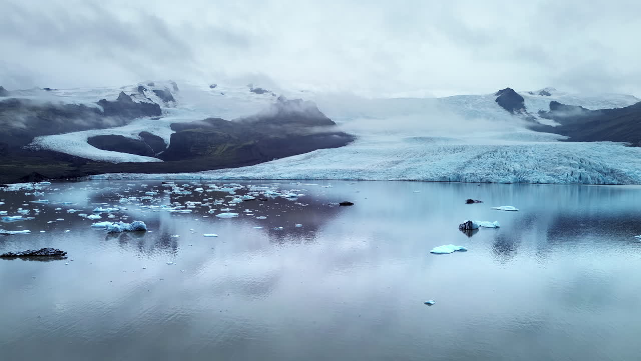 Luftfoto fra drone af Jökulsárlón gletsjerlagune i Sydøstisland. Flydende isbjerge driver hen over rolige, reflekterende farvande under Vatnajökull-iskappen