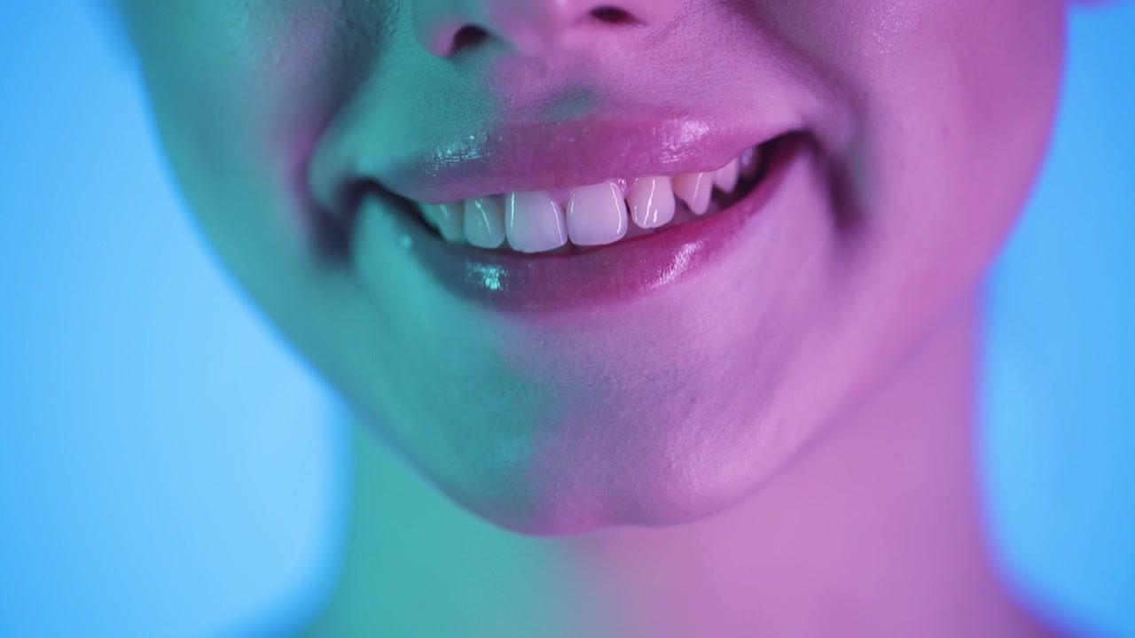 Slow motion 4K macro shot of a smiling woman licking a red heart-shaped lollipop, vibrant blue background with glowing pink and green neon light on her face, cinematic, playful, close-up scene