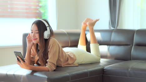 smiling young woman listens music with headphones lying on sofa barefoot