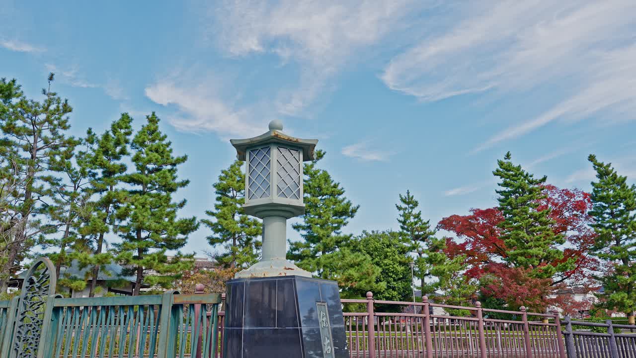 A traditional Japanese stone lantern on a bridge railing, set against a backdrop of pine and autumn trees under a cloudy sky