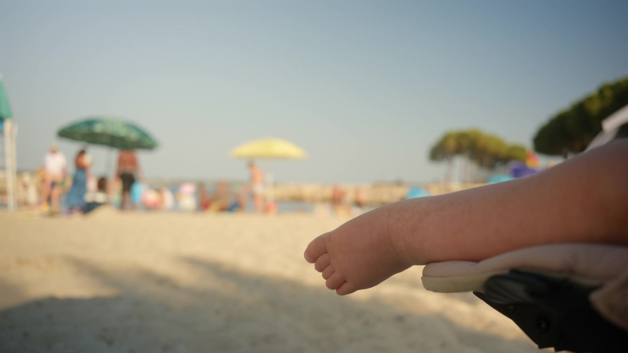 Il piedino di un bambino che riposa su un passeggino sulla spiaggia sabbiosa. Persone che si godono il sole e il mare sotto gli ombrelloni in uno sfondo sfocato.