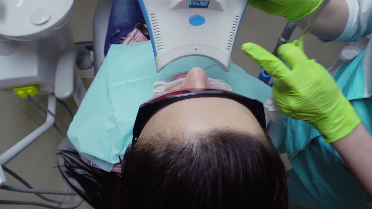 Young woman with an expander in mouth and red protective glasses getting UV whitening at the dentist's office by an ultra violet machine.
