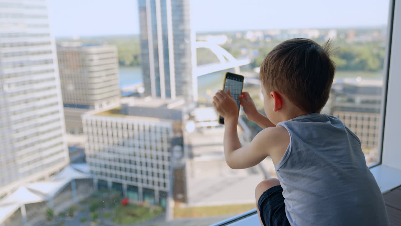 Boy playing with smartphone. A child in a gray tank top uses a smartphone while sitting by the window on a high balcony with city buildings below