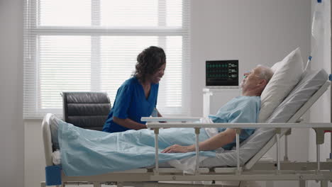 a dark-skinned female doctor is talking to an old male patient lying on a hospital bed discussing rehabilitation after recovery.