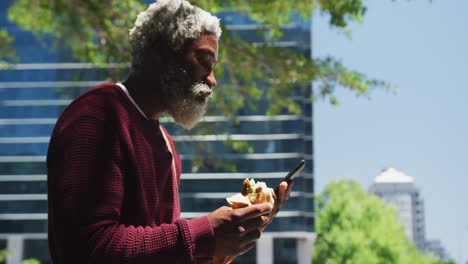 african american senior man having a snack and using smartphone at corporate park