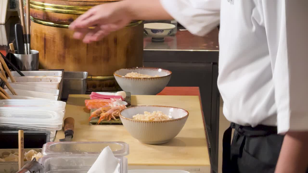 Ein professioneller Koch bereitet eine japanische Reisbowl mit Toppings an einem organisierten Restaurantschalter zu, statische Seitenansicht