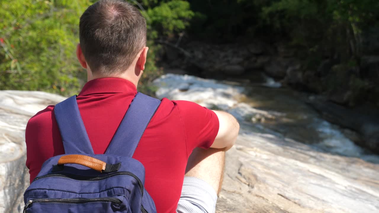 파란 배 을 입은 빨간색 폴로 셔츠 를 입은 남자 여행자 가 산 강 근처 에 앉아 있다