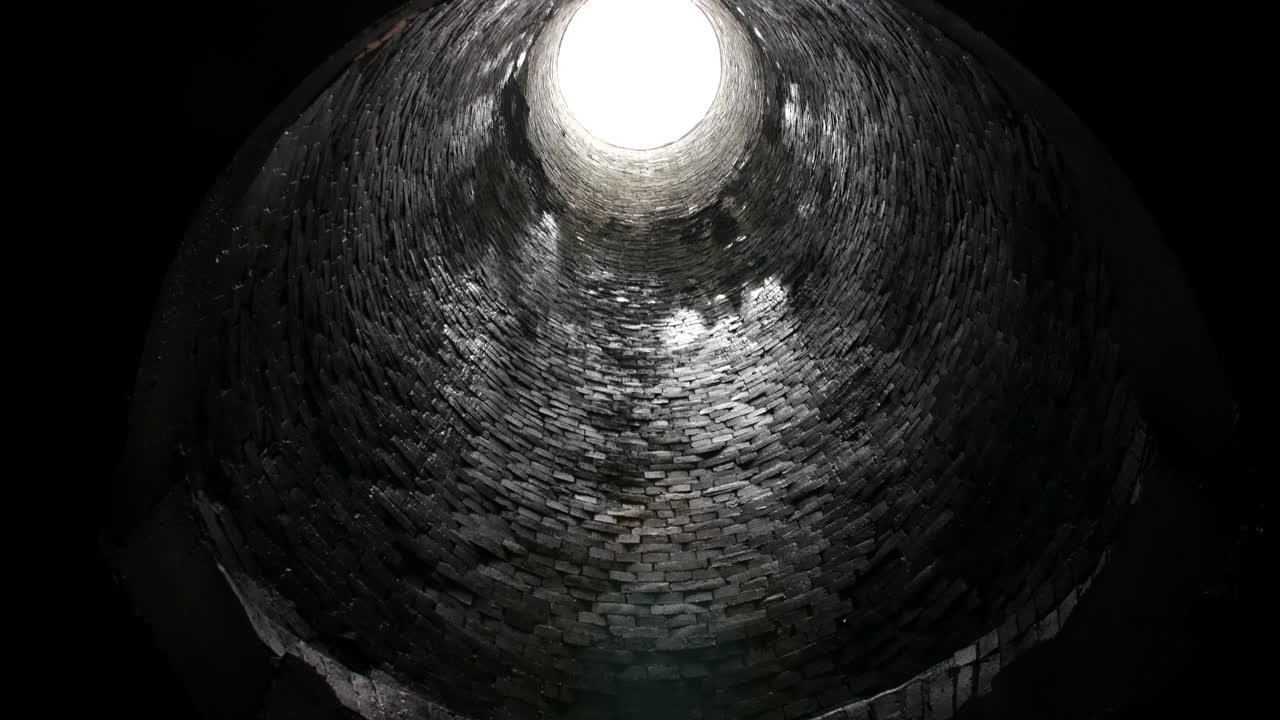 Looking up from inside a cylindrical brick tunnel, a bright circular opening at the top contrasts with the dark, damp walls below underground.