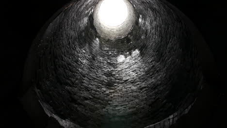 looking up from inside a cylindrical brick tunnel, a bright circular opening at the top contrasts with the dark, damp walls below underground.