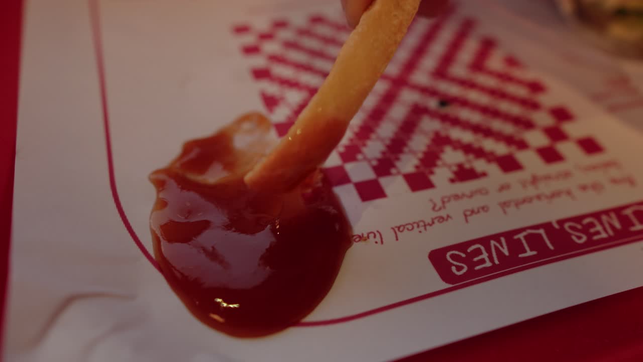 Nærbilde av hender som dypper en pommes frites i ketchup på papiret og tar den bort.