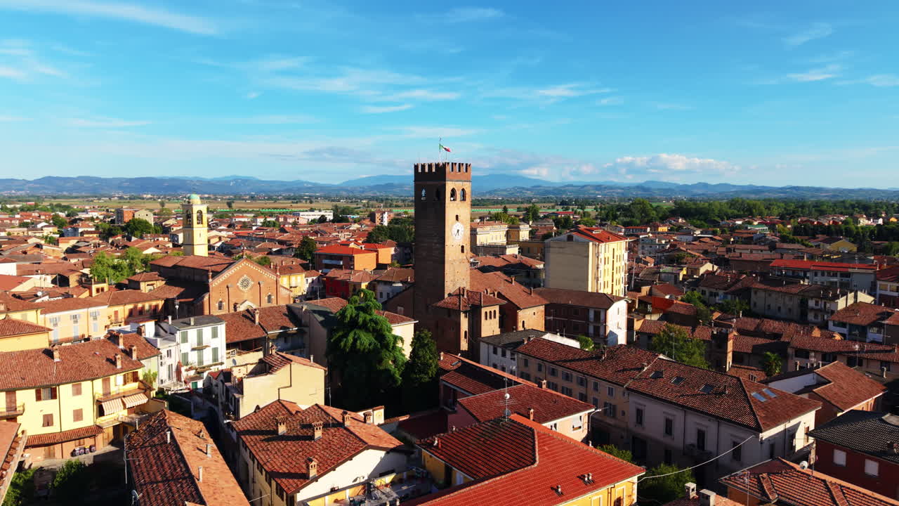 Ripresa aerea che si allontana da una torre medievale in pietra, rivelando i tetti illuminati dal sole di una storica città piemontese