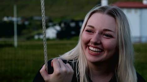 Happy-smiling-young-blonde-woman-swinging-under-big-tree-in-a-cold-green-spring-evening